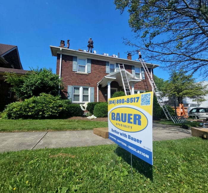 The image shows a house with a roof being installed by Bauer and a Bauer yard sign.