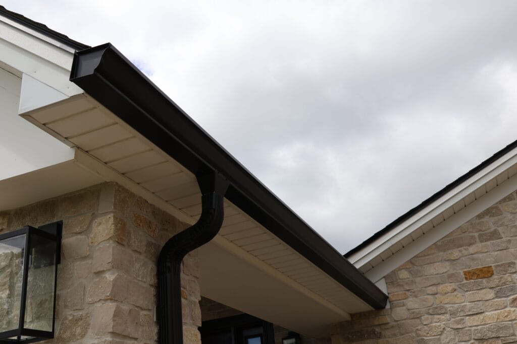 The image shows a close-up view of a newly installed black gutter system on a house with light-colored stone siding and white fascia.